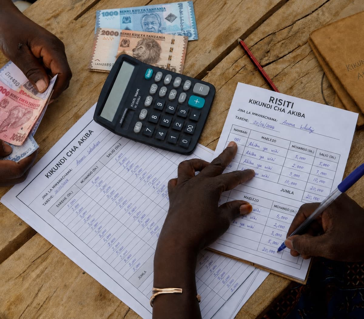 Overhead view of a savings group collection on a wooden table: one pair of hands counts Tanzanian shilling banknotes while another writes in a handwritten RISITI receipt, beside a desk calculator and a KIKUNDI CHA AKIBA ledger sheet ruled into TAREHE, MCHANGO and SALIO columns.